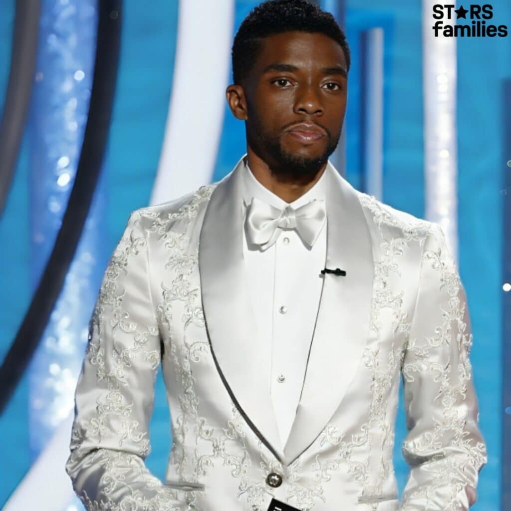 At a formal event, Chadwick Boseman poses in a white tuxedo with satin lapels and detailed embroidery, complemented by a white bow tie. He holds a black card or booklet, with his hands showcasing several rings and bracelets against a blue and white stage backdrop.