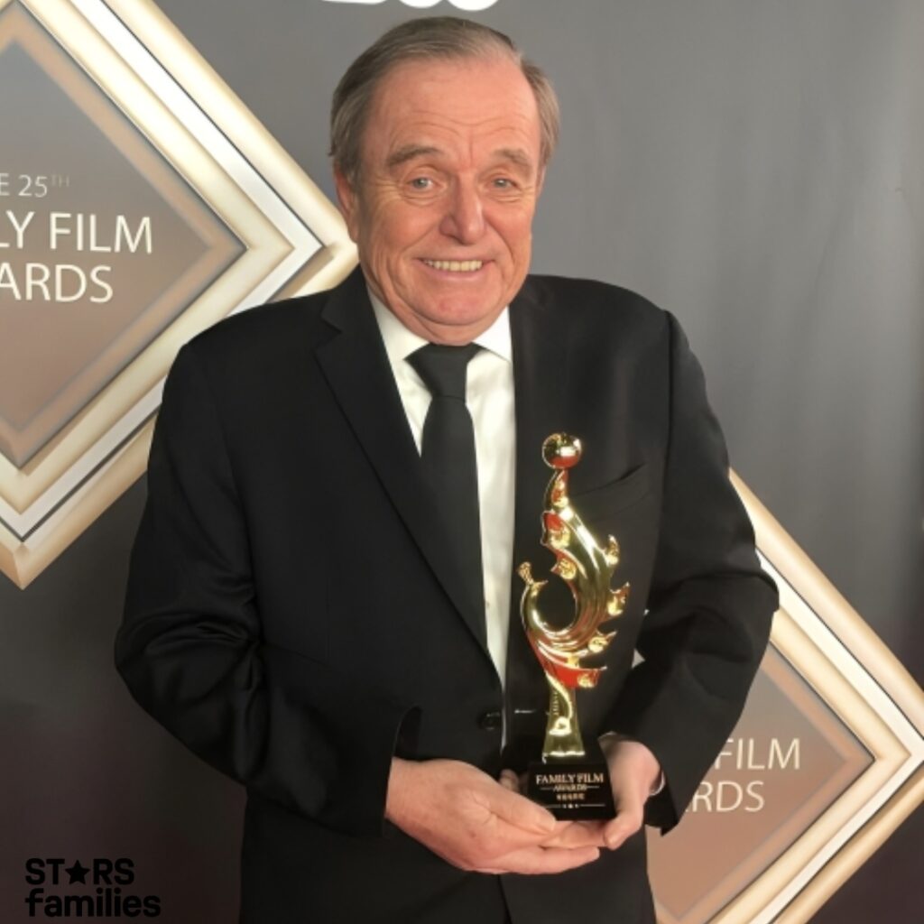 Jerry Mathers, wearing a black suit and tie, holds a gold trophy. He stands on a red carpet in front of a banner that reads "THE 25TH FAMILY FILM AWARDS" and "THE CW."