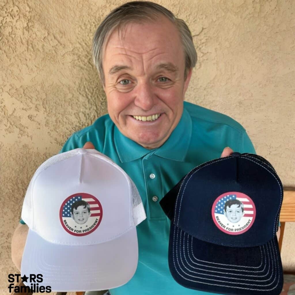 In front of a textured beige wall, Jerry Mathers, dressed in a teal polo shirt, holds a white and a black baseball cap. Both caps have a circular logo with an American flag, the text "SOMEONE FOR PRESIDENT."