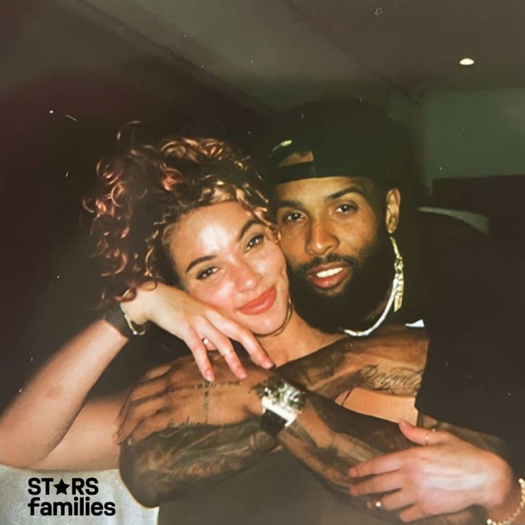 Lolo Wood embraces her husband in an indoor setting. Lolo has curly hair and is wearing a watch on her left wrist. Her husband has tattoos on his arms, is wearing a chain necklace, and has a watch on his right wrist.