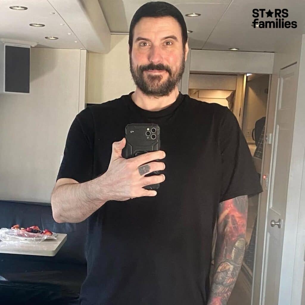 Benjamin Burnley in a selfie, smiling with a full beard and mustache, holding his phone in a mirror reflection. He wears a black t-shirt and shows a tattoo on his right arm.