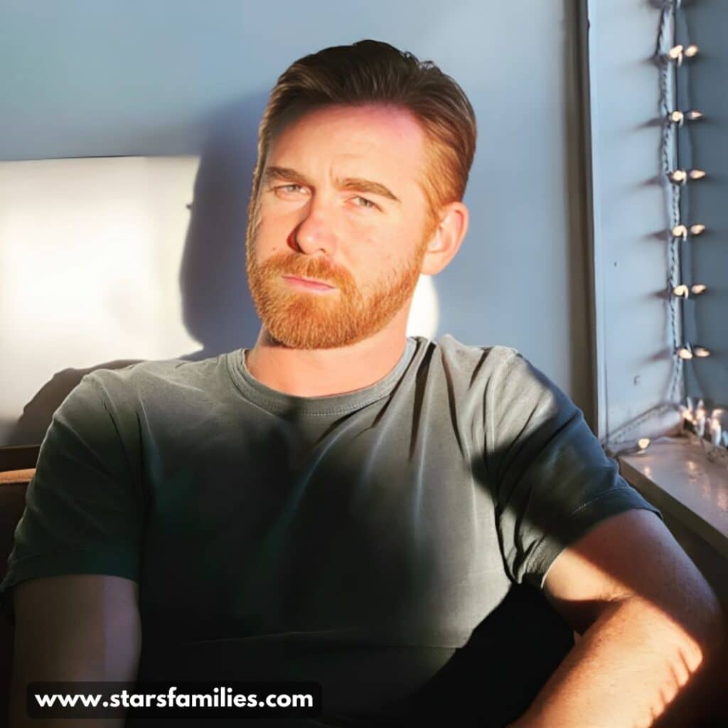 Andrew Santino, wearing a dark green t-shirt, sits indoors near a window. The sunlight streams through the window, creating shadows on his shirt and arm. String lights are visible along the window's edge, adding a cozy ambiance to the room.