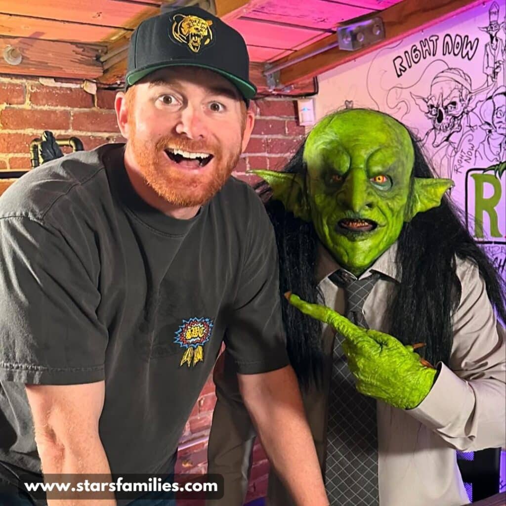 Andrew Santino, on the left, is wearing a black cap with a yellow logo and a dark t-shirt with a colorful graphic on the chest. Beside him, a person dressed in a costume with green makeup on the face and hands, a white shirt, a gray tie, and a black wig, points at Andrew.