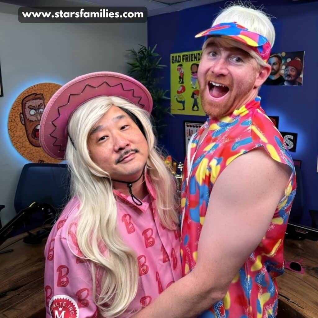 Andrew Santino, in a sleeveless multicolored shirt, matching headband, and short blonde wig, stands with a comedian wearing a pink hat, long blonde wig, and a pink outfit with the letter "B".