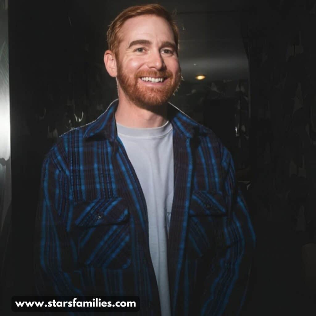 Andrew Santino stands in a dimly lit room, wearing a blue and black plaid shirt over a light-colored t-shirt.