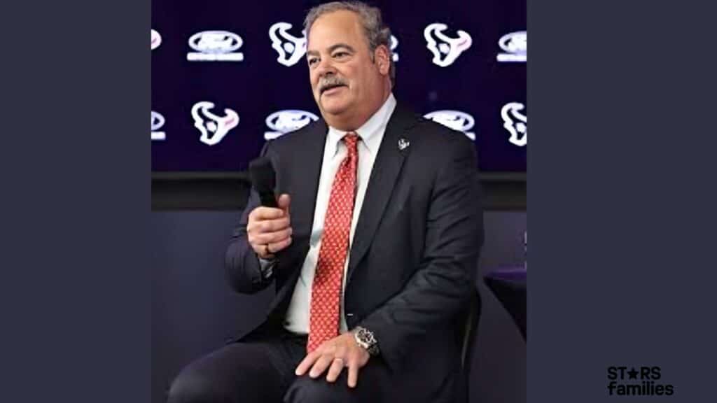 Cal McNair, wearing a dark suit with a white shirt and a patterned red tie, holds a microphone at a press conference