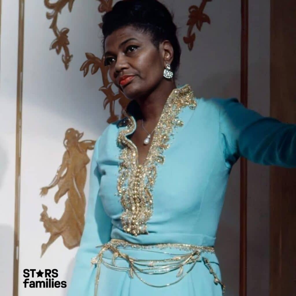 In a traditional and ornate light blue outfit adorned with gold embroidery, Pearl Bailey stands against a decorative wall with gold artistic designs.