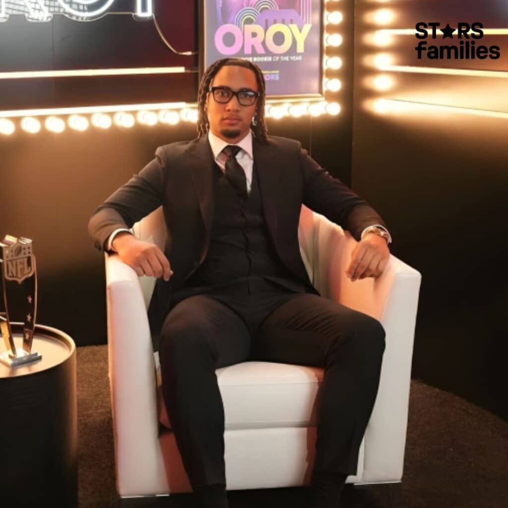 C.J. Stroud, dressed in a black suit, white shirt, and black tie, sits in a white armchair.