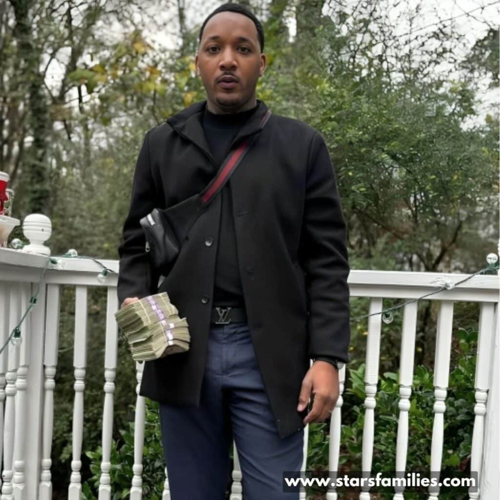 Jamal Morton stands on a porch or balcony with a white railing, holding a large stack of money in his left hand. He is wearing a black coat and dark pants.