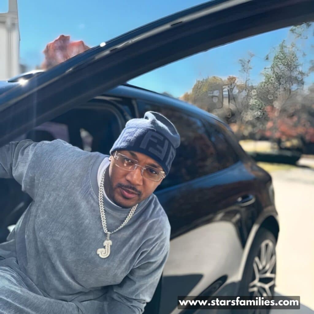 Jamal Morton, wearing a gray long-sleeve shirt, a gray beanie with a pattern, and a large chain necklace with a pendant in the shape of the letter "J," leans out of the open door of a black car.