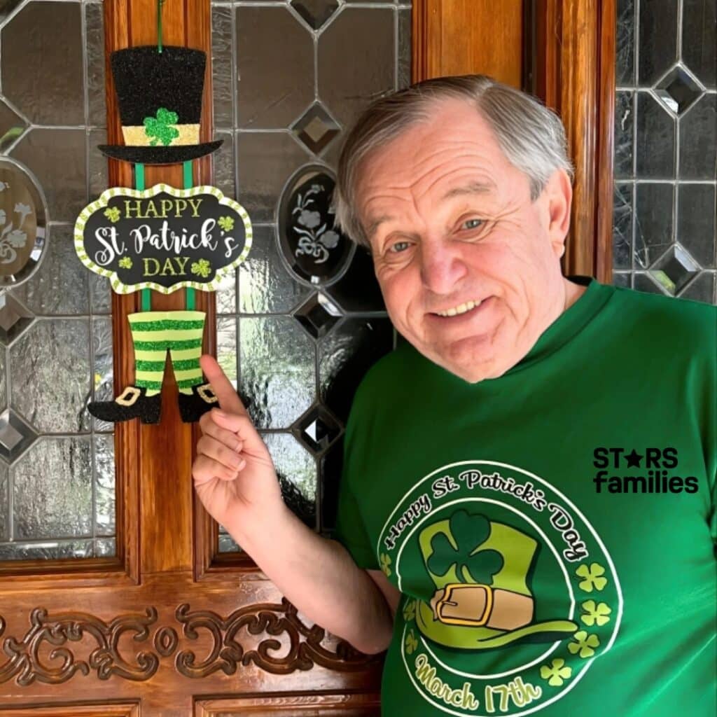 Jerry Mathers, wearing a green St. Patrick's Day-themed T-shirt with a shamrock, a leprechaun hat, and the text "Happy St. Patrick's Day" and "March 17th," stands in front of a wooden door with glass panels. He is pointing to a hanging decoration with a black top hat, green and black striped pants, and black shoes with gold buckles, which also reads "Happy St. Patrick's Day."