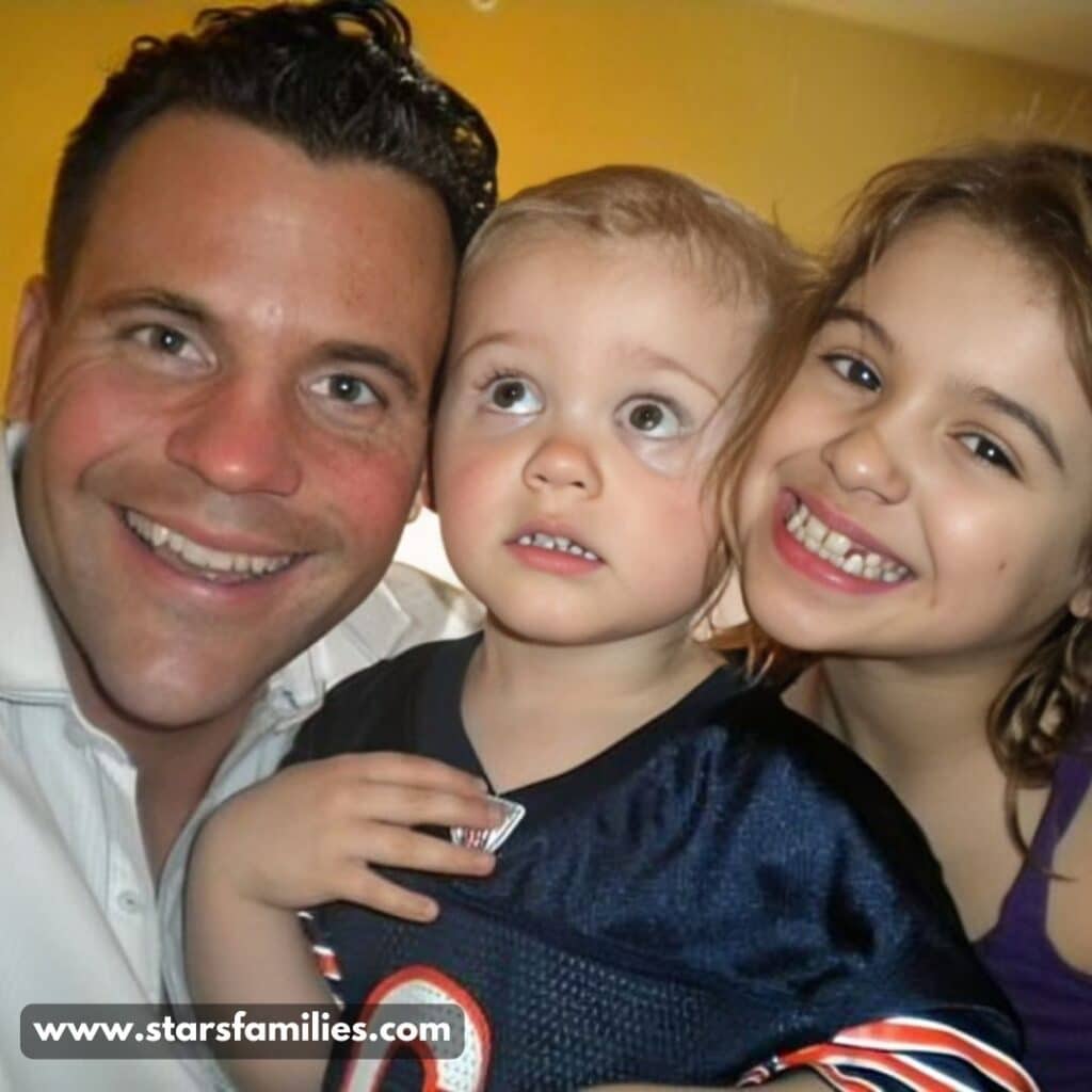 In an indoor environment with a yellowish wall, John Cerasani, wearing a white shirt, poses with his two children. One child is dressed in a dark sports jersey  and the other wears a purple top.