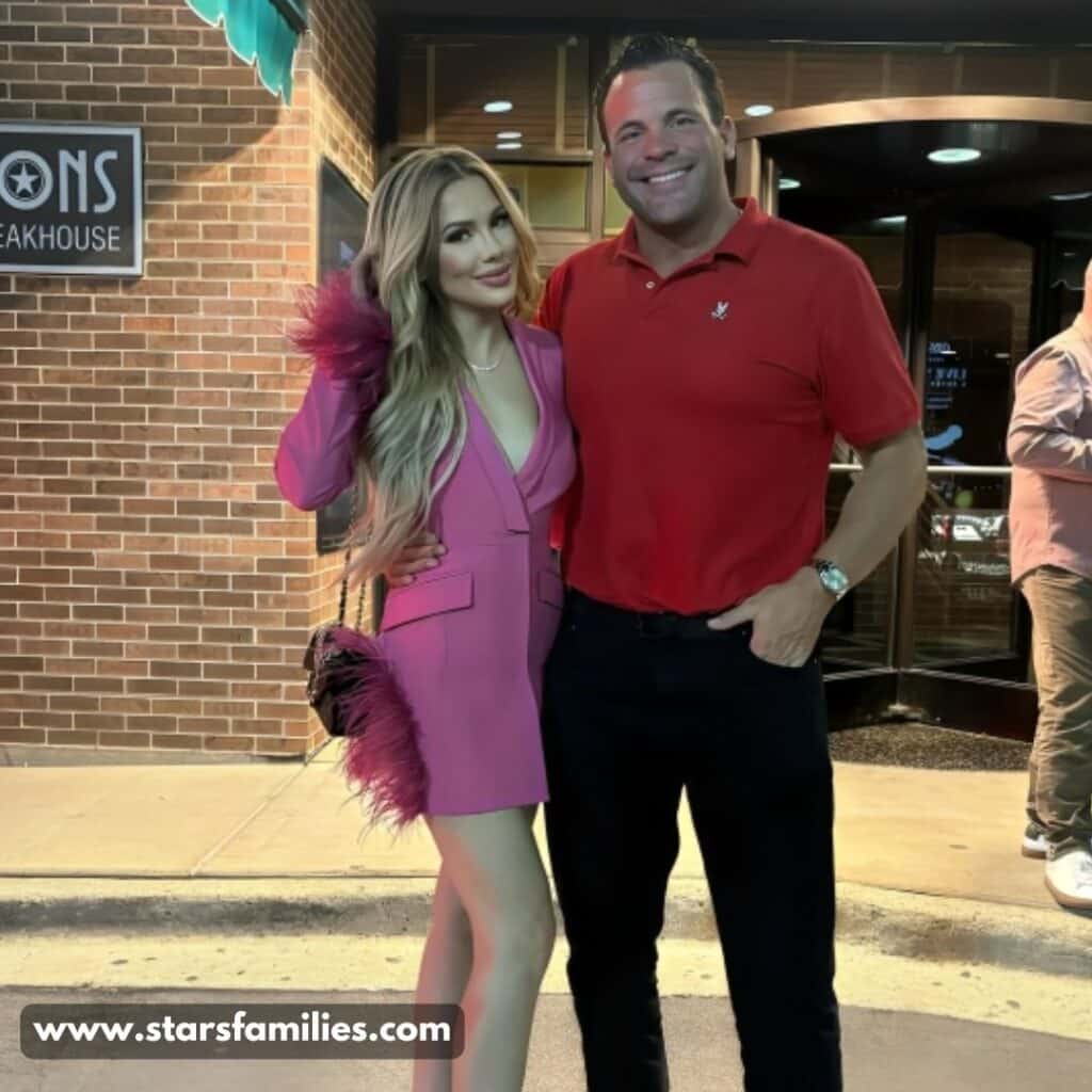 Andrew Santino, wearing a red polo shirt and dark pants, stands beside his wife, Sarah, who is dressed in a pink outfit and high heels, holding a feathered accessory. They are standing in front of Gibsons Bar & Steakhouse.