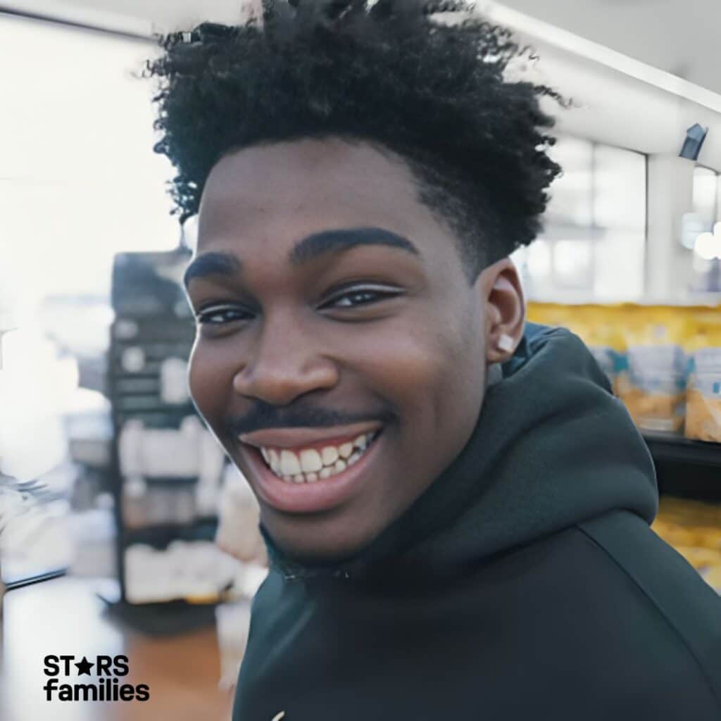 Kam Patterson, wearing a dark-colored hoodie, stands inside a store with shelves stocked with various items, including bags of snacks.