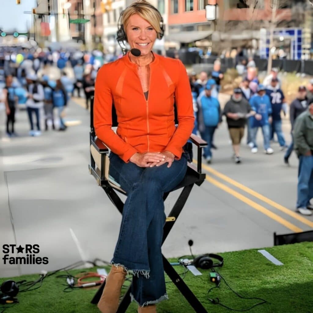 Karen Drew, dressed in a bright orange jacket, blue jeans with frayed hems, and tan ankle boots, is sitting on a director's chair on a platform covered with green artificial grass. She is wearing a headset with a microphone.