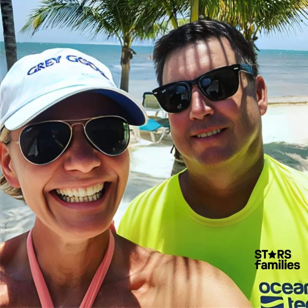 Karen Drew, wearing a white cap with the text "GREY GOOSE" and a pink strap around her neck, stands beside her husband, Jason Drew, who is wearing a bright yellow shirt with the text "ocean." They are on a sandy beach with palm trees, beach chairs, and the blue ocean in the background, suggesting a tropical vacation setting.