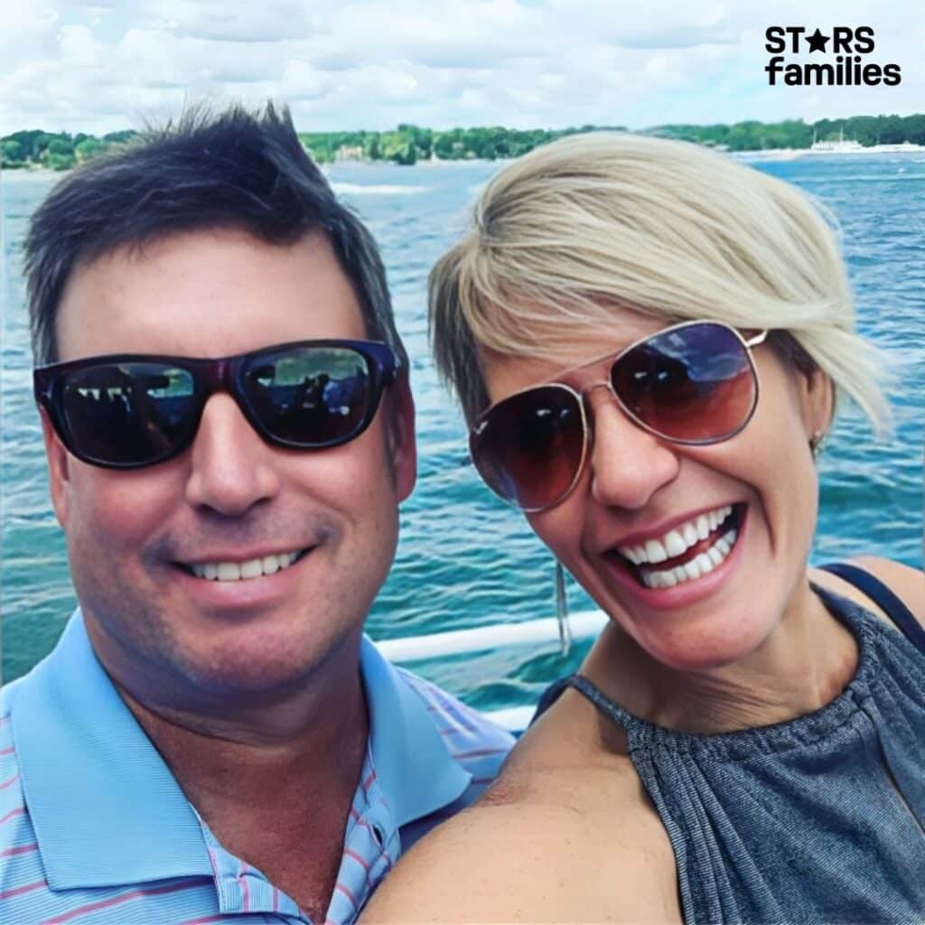 Karen Drew, wearing a light blue collared shirt with stripes, stands beside her husband, Jason Drew, who is dressed in a sleeveless top. They are standing near a waterfront with a calm body of water and a distant shoreline in the background.