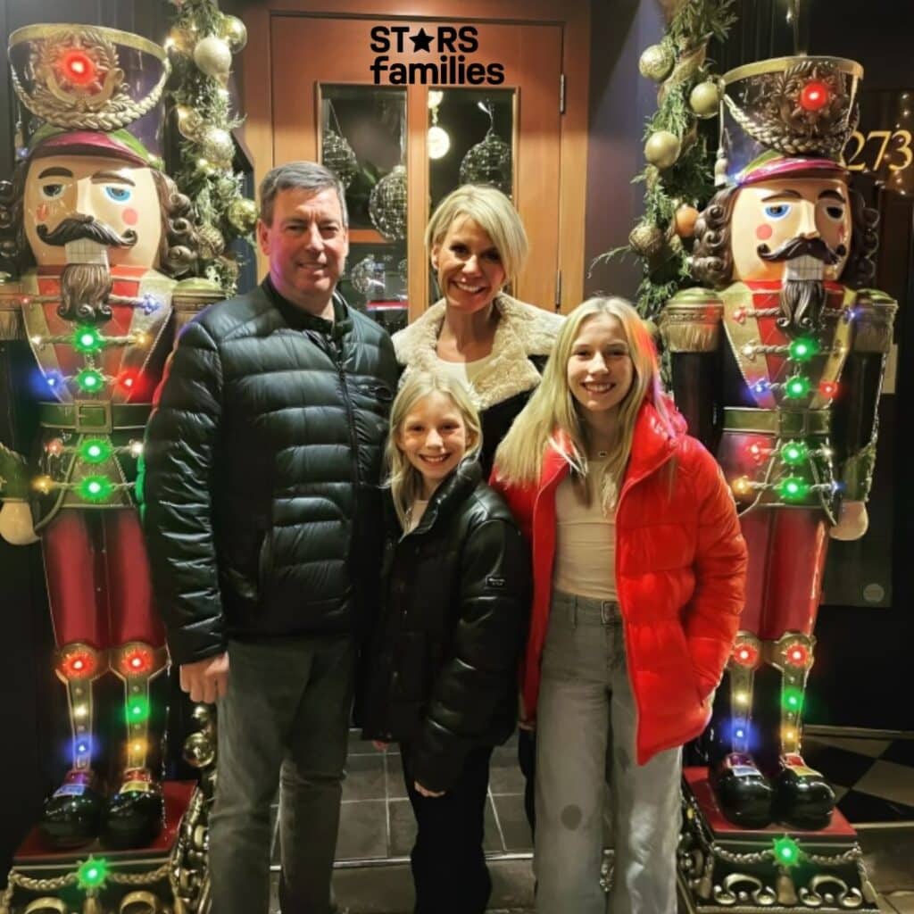 Karen Drew, dressed in a red coat, stands beside her husband, Jason Drew, who is wearing a black jacket. They are accompanied by their two children, both in winter clothing, standing in front of a festively decorated entrance. The entrance features two large nutcracker statues adorned with colorful lights, garlands, and spherical ornaments, suggesting a Christmas holiday season.