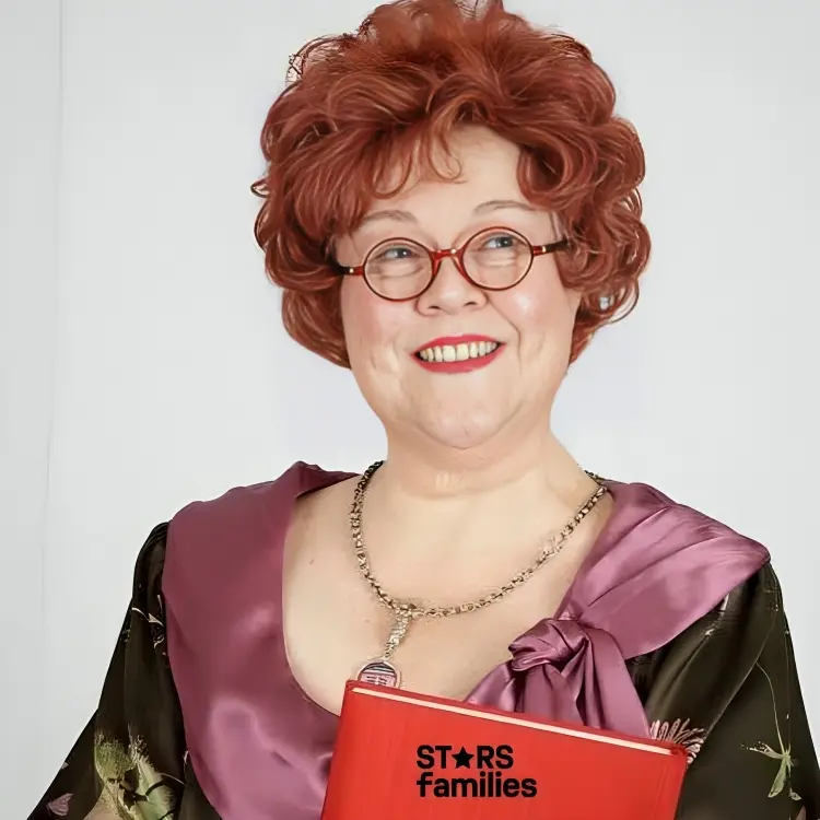 Kathy Kinney, with curly reddish hair, wearing a colorful outfit with a purple and green floral pattern and a purple satin-like fabric around the shoulders. She holds a red book and wears a necklace with a pendant. The background is plain white.
