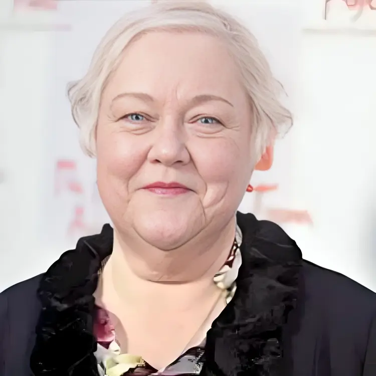 Kathy Kinney, dressed in a colorful and patterned scarf with shades of pink, green, and white, stands in front of a step-and-repeat banner at an event.
