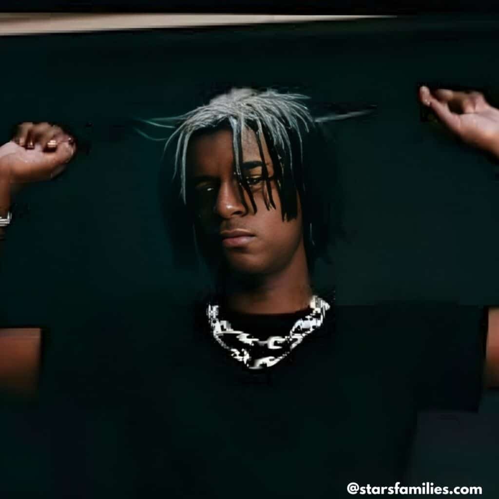 Ken Carson, with light-colored hair, raises both arms while wearing a black shirt and a chunky chain necklace.