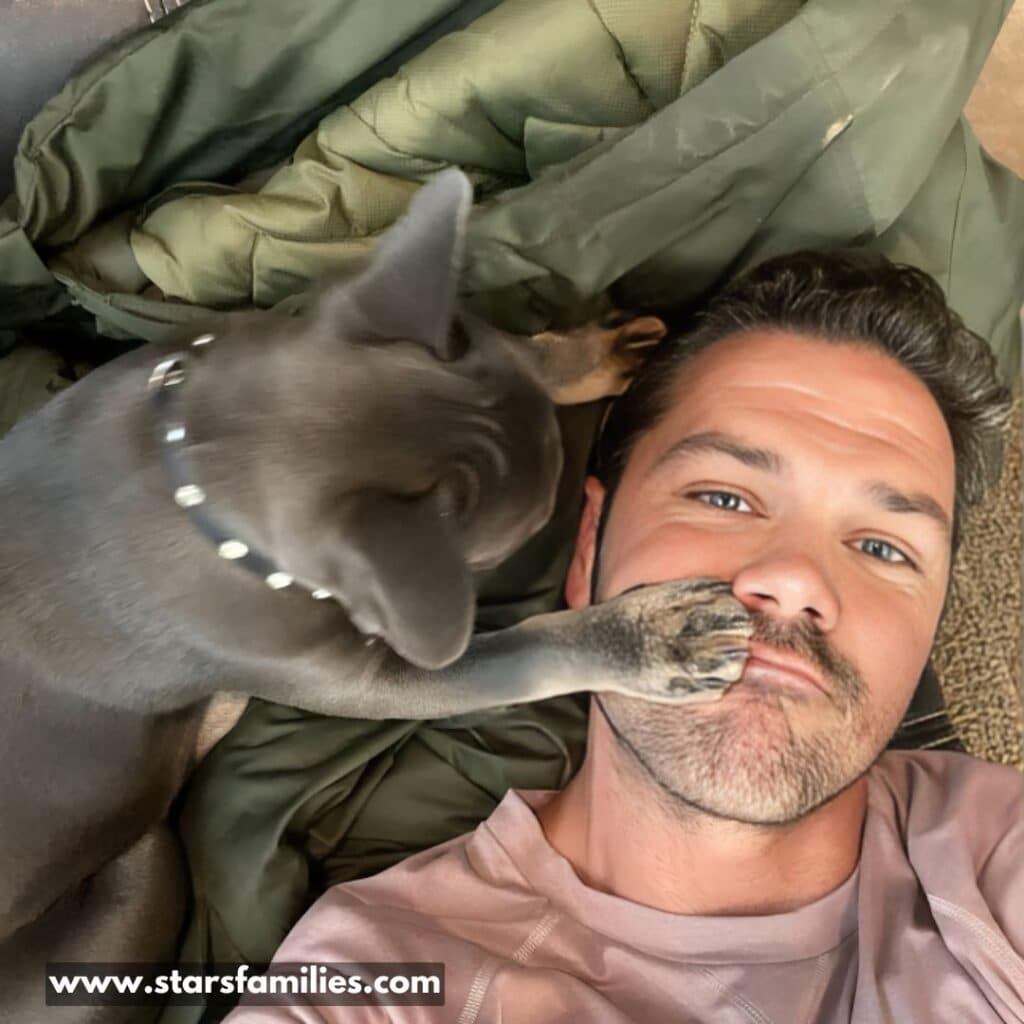 Ryan Paevey lies down with a dog. The dog, which has a gray or blue-colored coat and wears a studded collar, is positioned close to his head. Ryan is wearing a light-colored shirt, and there is a green quilt or blanket underneath them