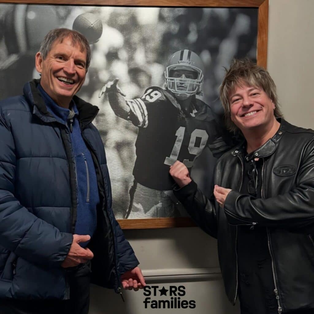 In front of a framed black-and-white photograph of a football player with the number 19, Paul Sidoti, dressed in a blue jacket, smiles and points towards the photo. Another person in a black leather jacket stands beside him, also pointing at the photograph.