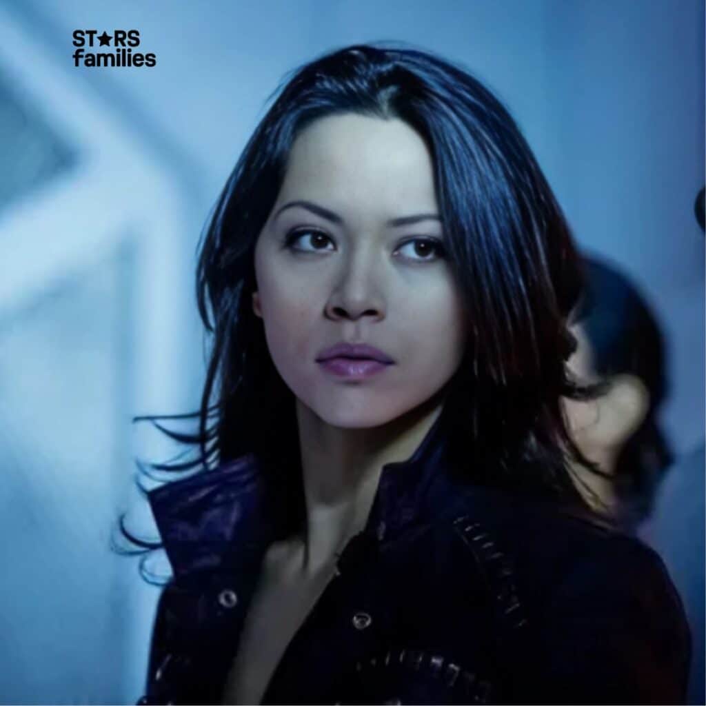 In an indoor setting with a blue tint, Melissa O'Neil, with long dark hair, is dressed in a dark-colored jacket.