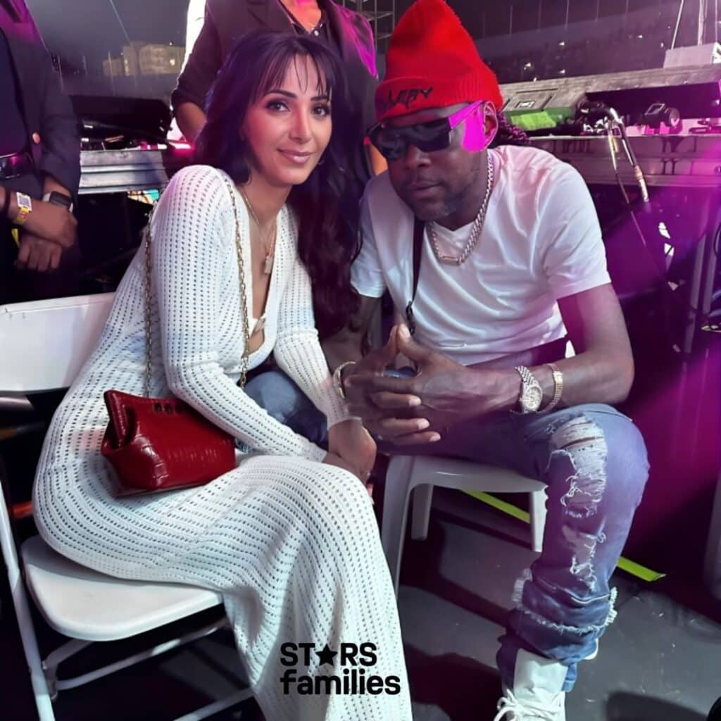 Vybz Kartel, wearing a red beanie, a white t-shirt, ripped jeans, and white sneakers, sits next to Sidem Ozturk, who is dressed in a long, white, knitted dress and holding a red handbag. They are seated on white chairs in a backstage or VIP area at an event.