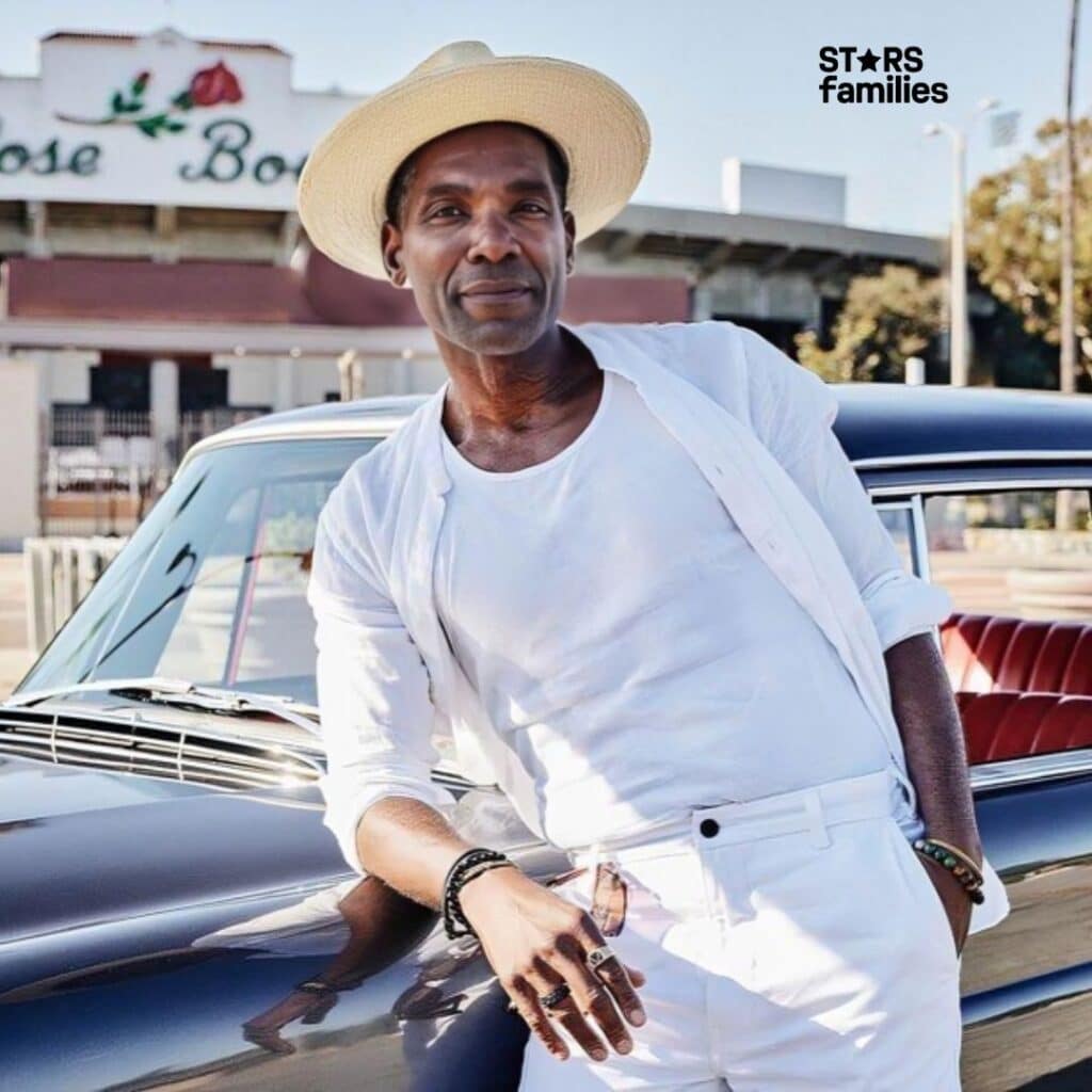 Ray Campbell, wearing a white outfit including a white hat, white shirt, and white pants, leans casually against a vintage car with a dark exterior and red interior. The background features a building with a sign that reads "Rose Bowl," trees, and a clear sky.