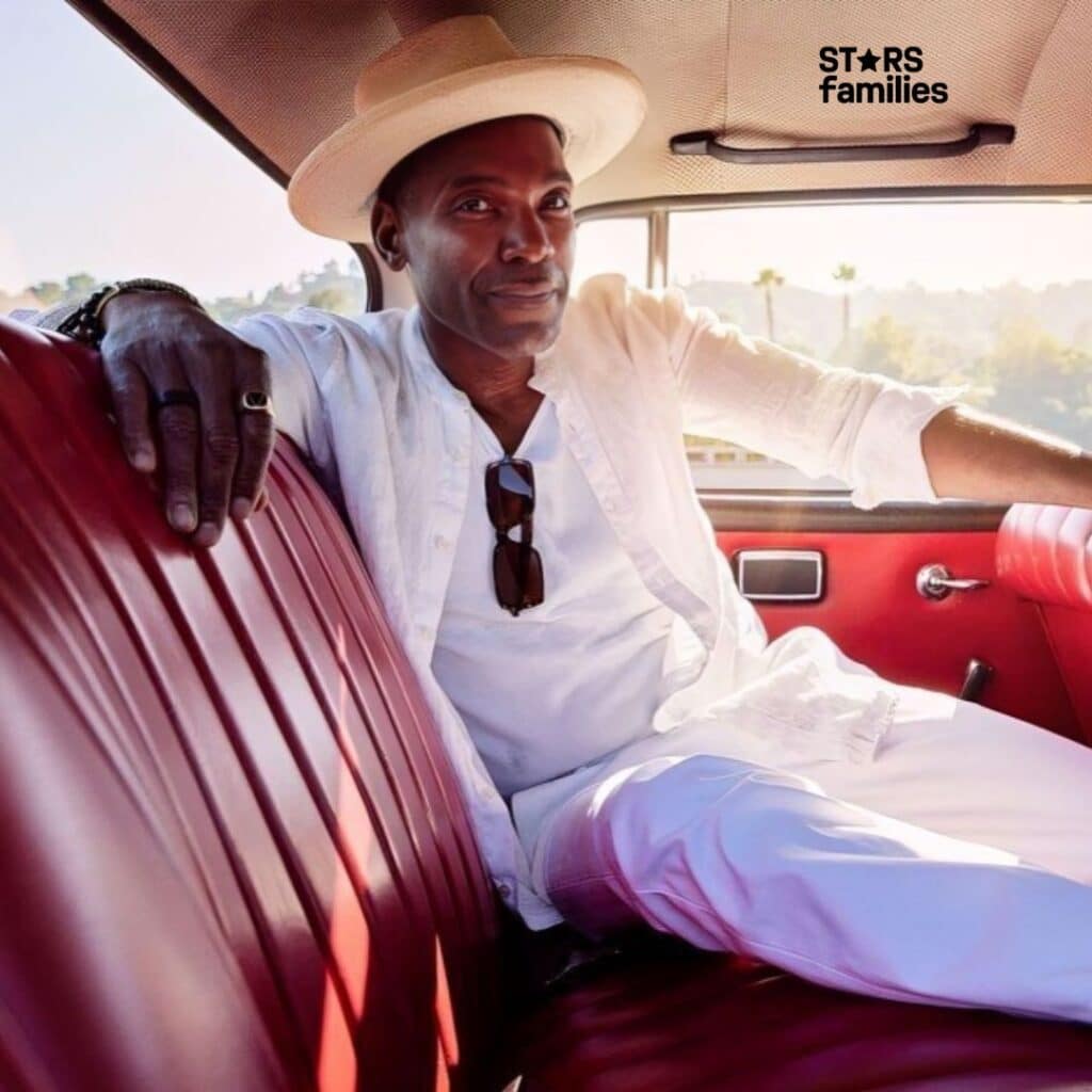 Ray Campbell, wearing a white shirt, white pants, and a white hat, sits in the back seat of a car with red leather interior. He has sunglasses hanging from his shirt and rests one arm on the back of the seat. Through the window, trees and hills are visible.