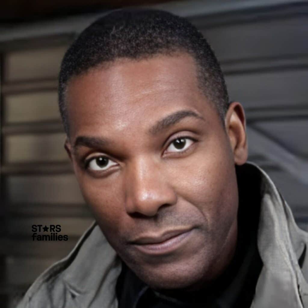 Ray Campbell, wearing a light-colored utility jacket over a dark shirt, stands in front of an indoor setting with metallic or industrial elements like a corrugated metal wall or door.