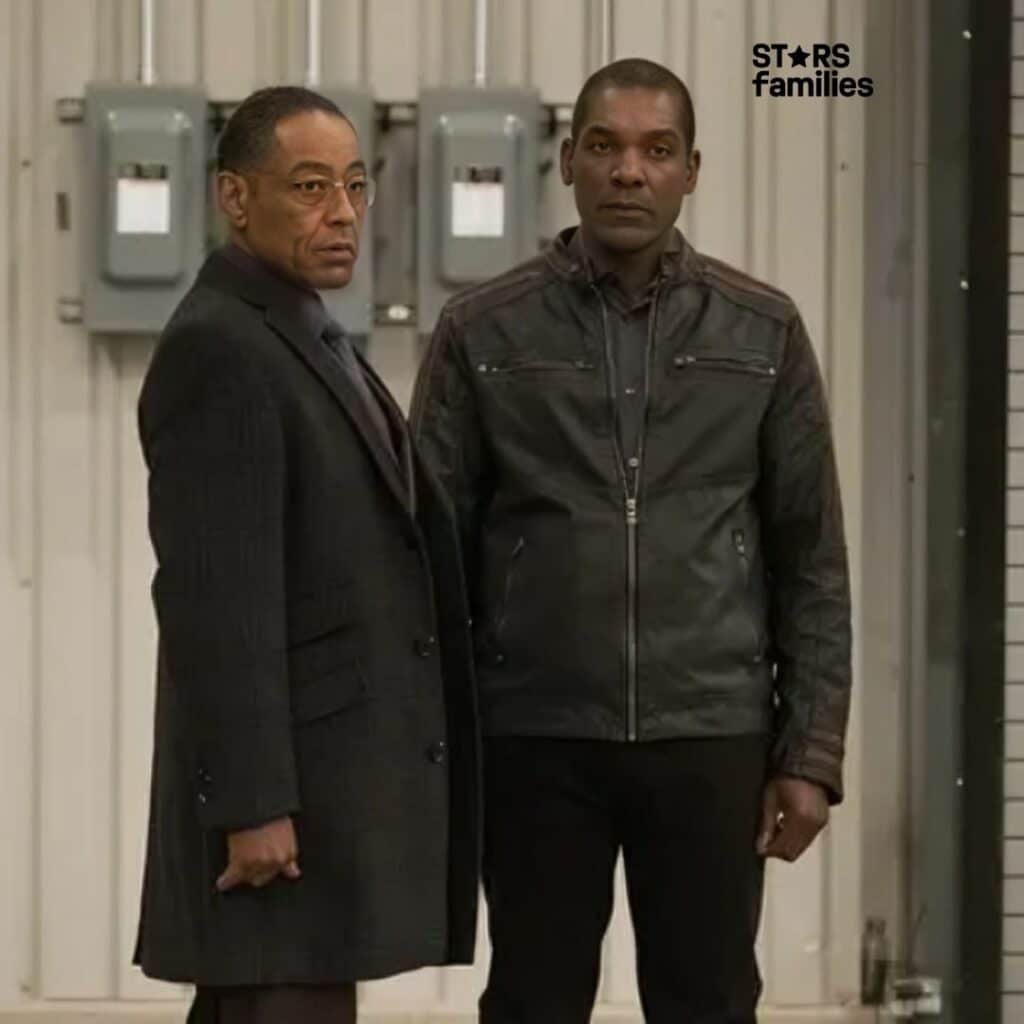 Ray Campbell, wearing a dark overcoat, stands indoors beside Giancarlo Esposito wearing a dark leather jacket. They are in front of a wall with electrical panels and a fire alarm, suggesting an industrial or utility area, possibly a warehouse or maintenance facility.