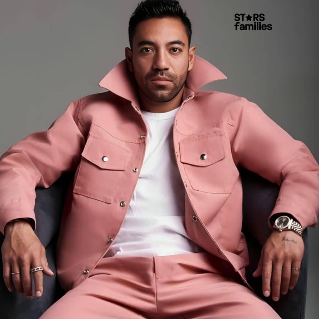 Marco Fabián, wearing a pink jacket with silver buttons and matching pink pants, sits on a chair. He has a white shirt underneath the jacket and is accessorized with a watch on his left wrist and two rings on his right hand. The background is plain.