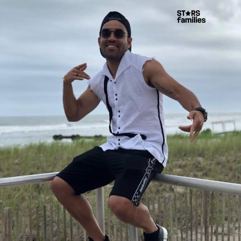 Marco Fabián, wearing a sleeveless white shirt with black accents and black shorts with white text and designs on the side, poses outdoors near a beach. The background features a grassy area with a wooden fence and the ocean with waves visible in the distance. The sky is overcast.