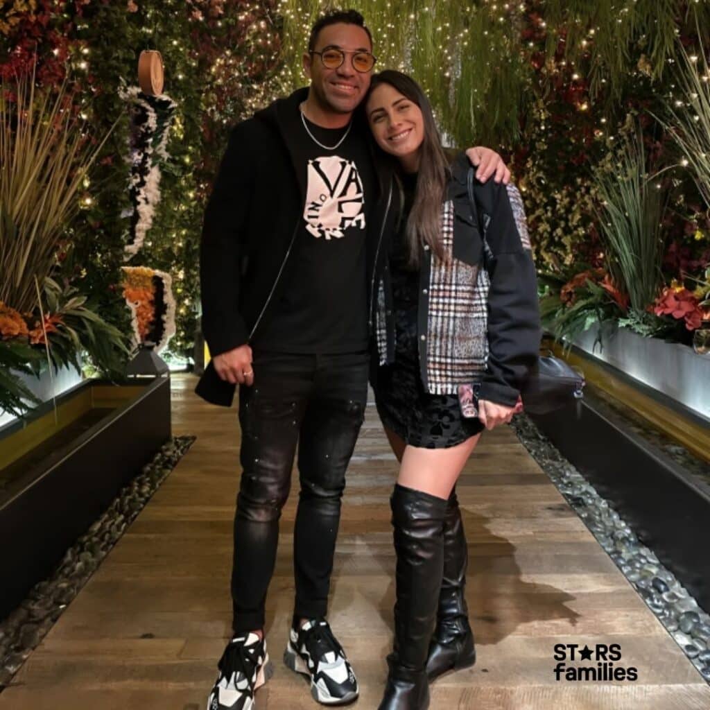 Marco Fabián, wearing a black jacket, black t-shirt with a white graphic, black jeans, and black and white sneakers, stands next to his girlfriend. She is dressed in a black and white checkered jacket, a black dress, and black knee-high boots. They are standing together in a well-decorated area with lush greenery and numerous small lights, creating a festive and enchanting atmosphere. The setting appears to be an indoor garden with a wooden pathway and water features on either side.