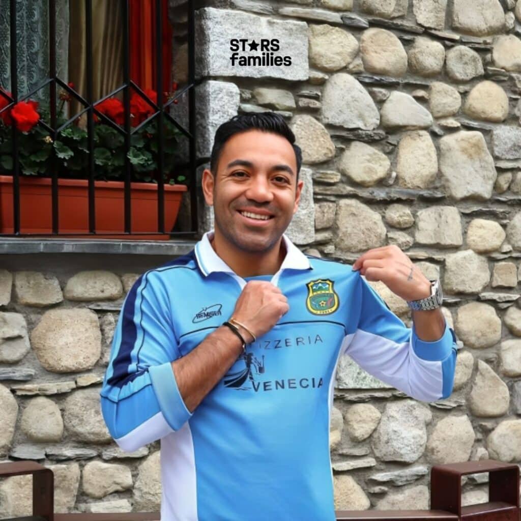 Marco Fabián, wearing a light blue sports jersey with the text "PIZZERIA VENEZIA" on the front and a logo on the left chest, stands in front of a stone wall with a window that has a flower box containing red flowers. He is holding the collar of the jersey with both hands. The background includes a stone wall and a window with a red frame and black bars.