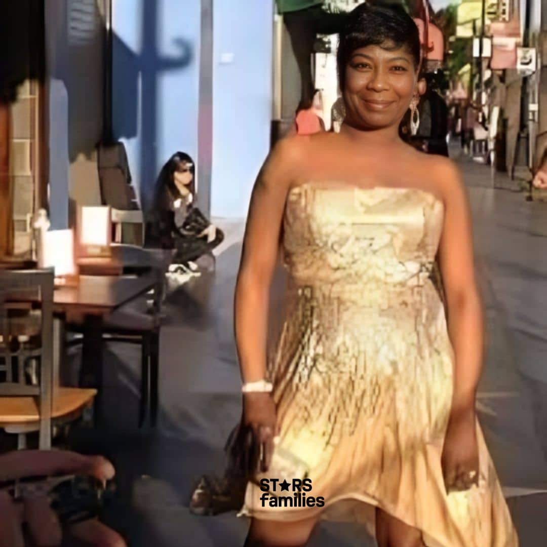 Sweet Brown, wearing a strapless, asymmetrical dress with a gradient effect transitioning from light to dark shades, walks on a sidewalk. She is holding a small clutch bag and wearing high-heeled shoes. The background includes other people, some sitting at tables outside a café or restaurant, and others walking. The sidewalk has stars embedded in it, similar to the Hollywood Walk of Fame. The image is taken in daylight, with shadows indicating the direction of the sunlight.