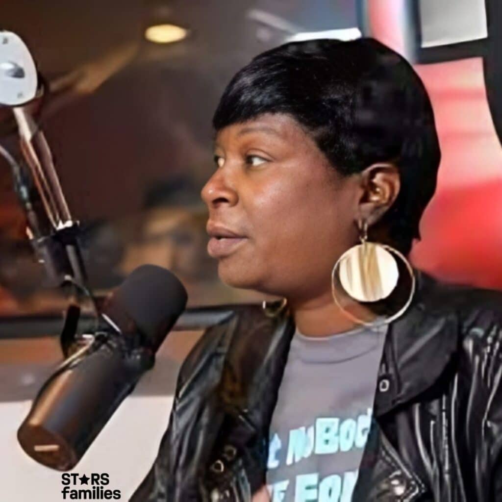 Sweet Brown, wearing a black leather jacket and large hoop earrings, speaks into a microphone in a radio station or recording studio. The background features a logo with the number "97" prominently displayed, indicating the station's frequency or branding. The setting suggests an interview or live broadcast.