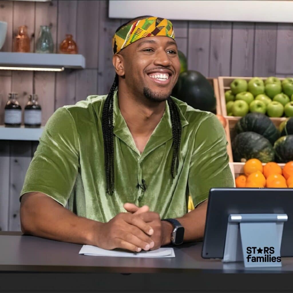 In an indoor setting, Darnell Ferguson, dressed in a green velvet shirt and a colorful headband, sits at a desk with hands clasped together.The background includes shelves with jars, bottles, and a variety of fruits and vegetables, such as green apples, oranges, and squash or pumpkins.