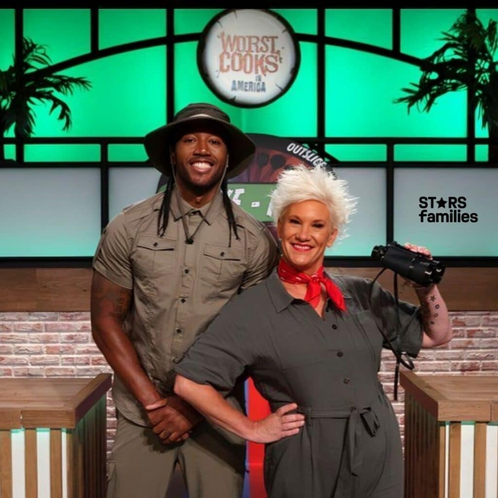 Darnell Ferguson, wearing a khaki-colored outfit with a wide-brimmed hat, stands next to the lady, who is dressed in a gray jumpsuit with a red bandana around her neck and holding binoculars. They are standing in front of a backdrop with the logo "Worst Cooks in America." The backdrop features a green and white color scheme with some plants.