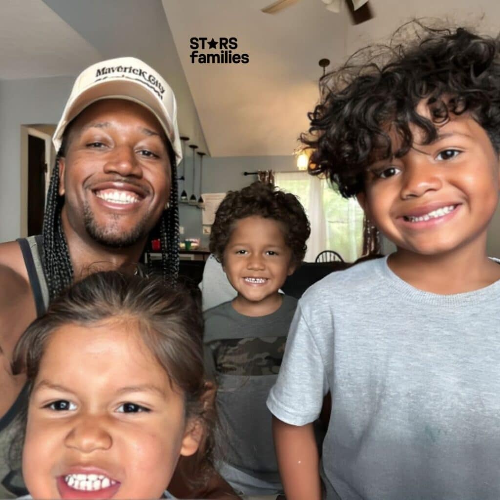 Darnell Ferguson, wearing a black t-shirt with a graphic design, stands with 3 children in front of him. He is wearing a white cap with the text "Maverick City" on it.