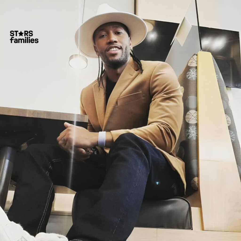 Darnell Ferguson, wearing a light-colored hat, a tan blazer, dark jeans, and white shoes, sits on a bench or seat in a modern indoor setting. The background includes a table, a hanging light fixture, and a wall with a patterned design.