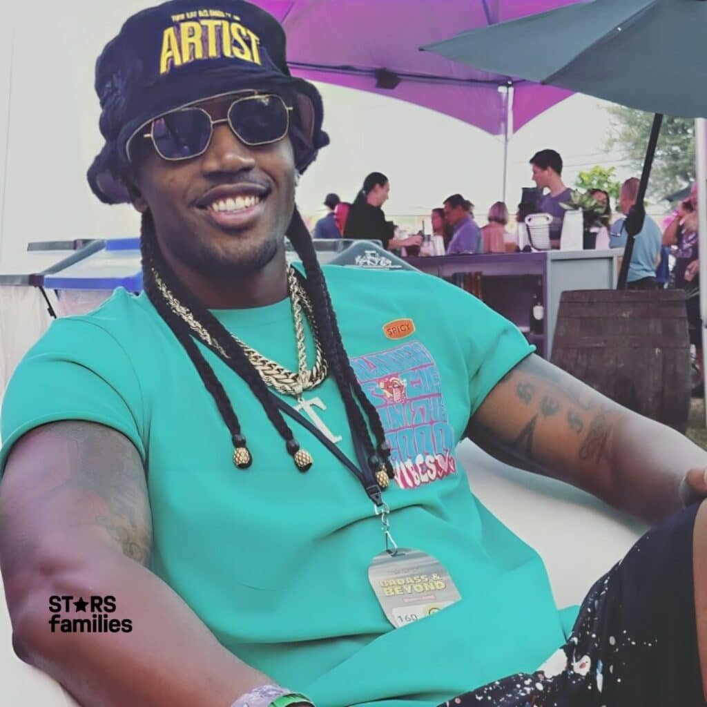 Darnell Ferguson, wearing a black hat with the word "ARTIST" in yellow letters, a turquoise shirt with a colorful graphic design and the word "SPICY" on a small orange label, and multiple gold chains around the neck, sits outdoors.