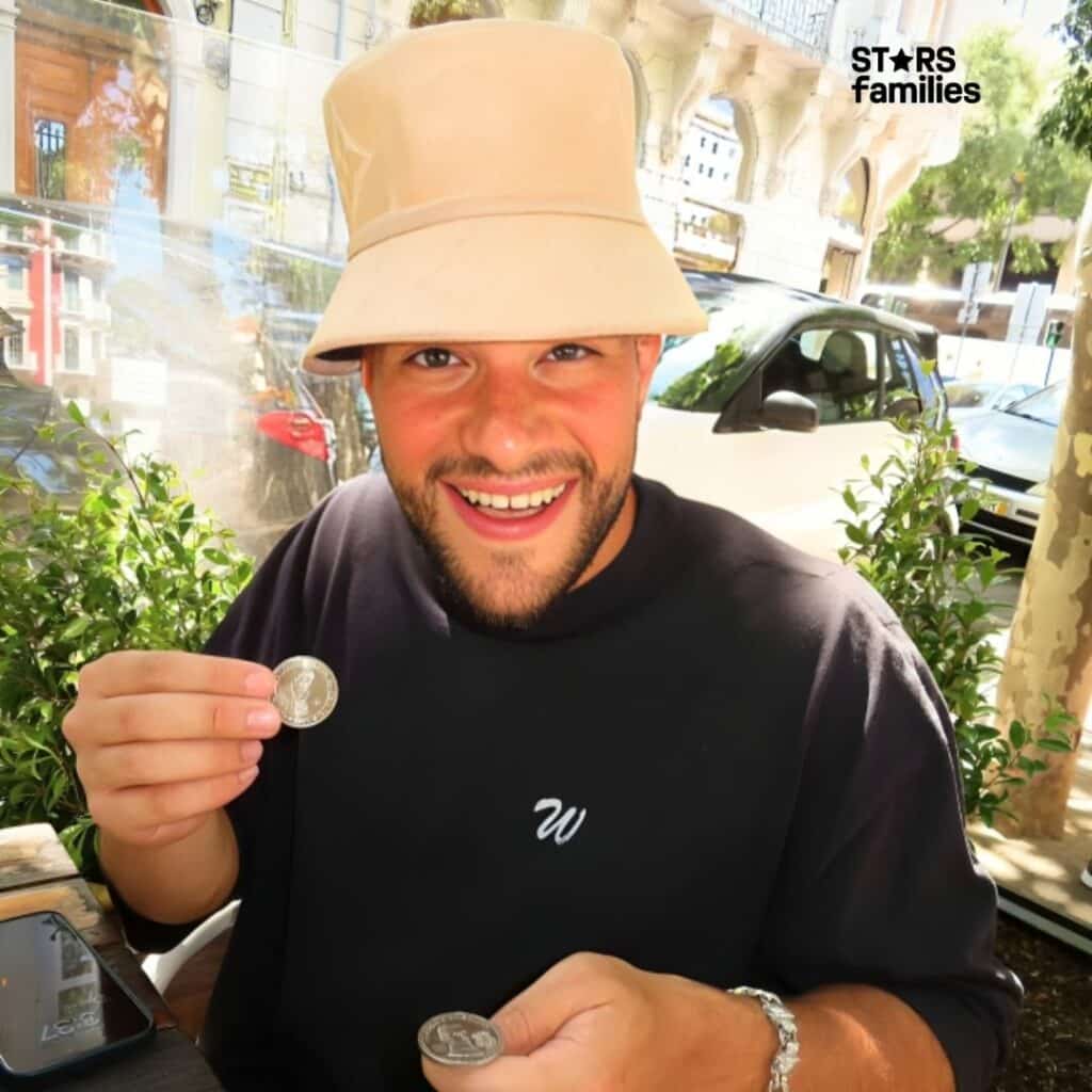 Moses the Jeweler, wearing a beige bucket hat and a black shirt with a white "W" logo, sits outdoors holding two coins. He is holding one coin in each hand. A smartphone is placed on the table in front of him. The background includes greenery, a white car, and buildings.