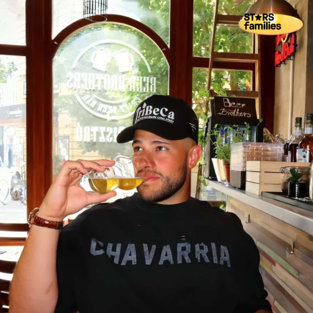 Moses the Jeweler, wearing a black cap with the text "TriBeCa" and a black shirt with the text "CHAVARRIA," sits casually in a bar or restaurant setting. He is holding a glass of beer near his mouth. The background includes a window with the text "BEER BROTHERS" and "BEER FACTORY," along with an image of two beer mugs clinking together. There is also a sign on a shelf that reads "Beer Brothers," with various bottles and plants visible.