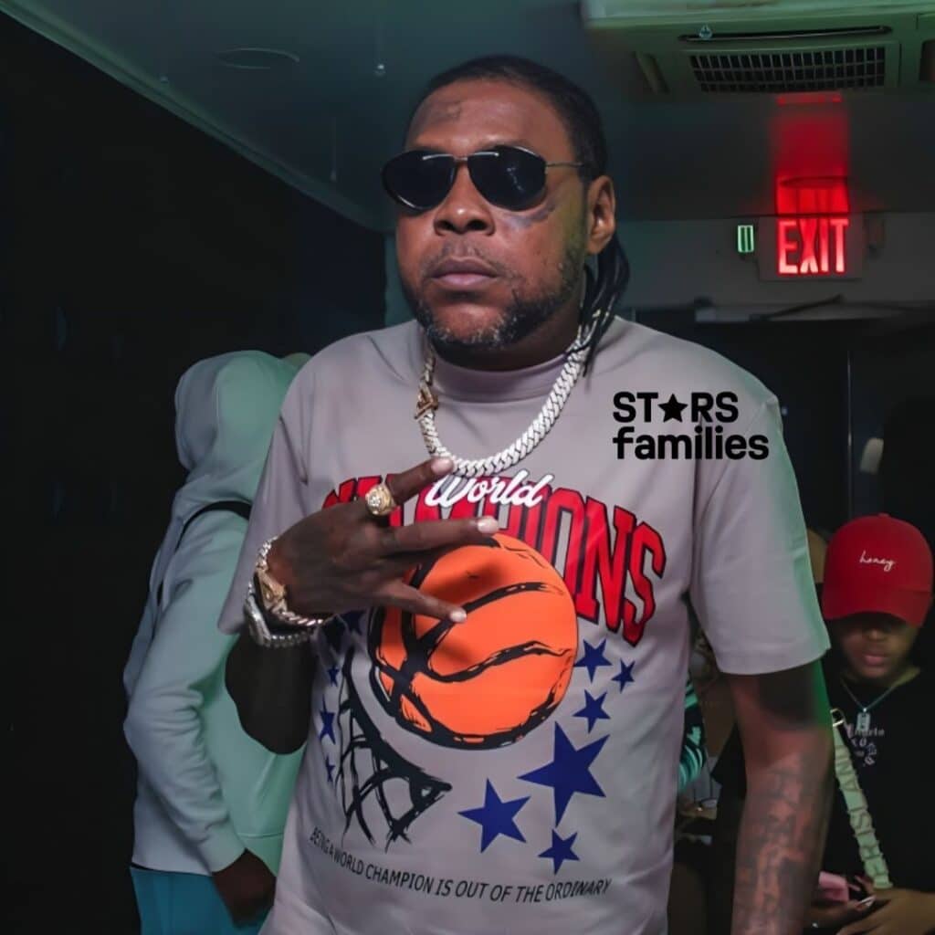 Vybz Kartel, wearing a gray t-shirt with a basketball graphic and the text "World Champions" in red letters, stands confidently. He has a thick silver chain necklace and a ring on his finger.