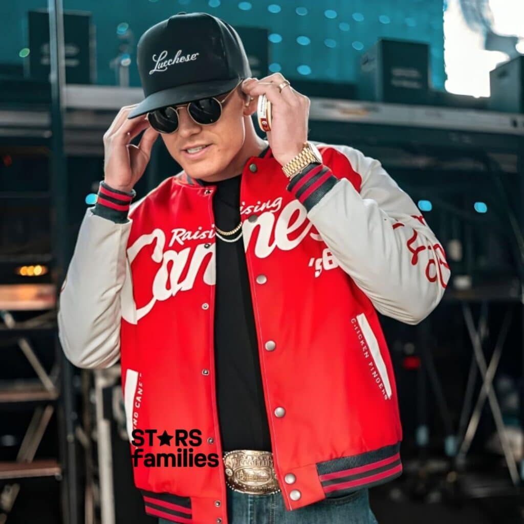 In a concert or stage setting, Parker McCollum is dressed in a red and white jacket with "Raising Cane's" written on it. He wears a black cap with "Lucchese," a gold watch, a gold chain, and a large belt buckle.