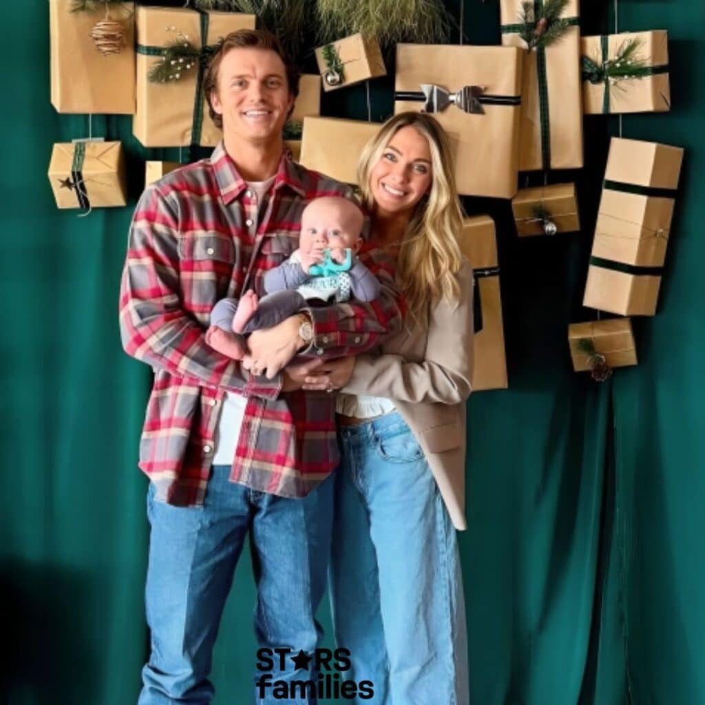 Parker McCollum, wearing a red and gray plaid shirt and blue jeans, stands with his wife, who is dressed in a beige blazer and light blue jeans, and their son, who is wearing a blue outfit. They are standing in front of a green backdrop decorated with wrapped gifts in brown paper with green and black ribbons, some adorned with small pine branches and ornaments.