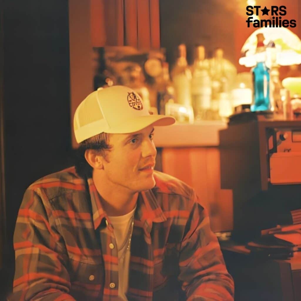 Parker McCollum, wearing a white cap with a logo, a plaid shirt, and a white t-shirt underneath, stands in a dimly lit room with various bottles and a lamp on a shelf in the background.